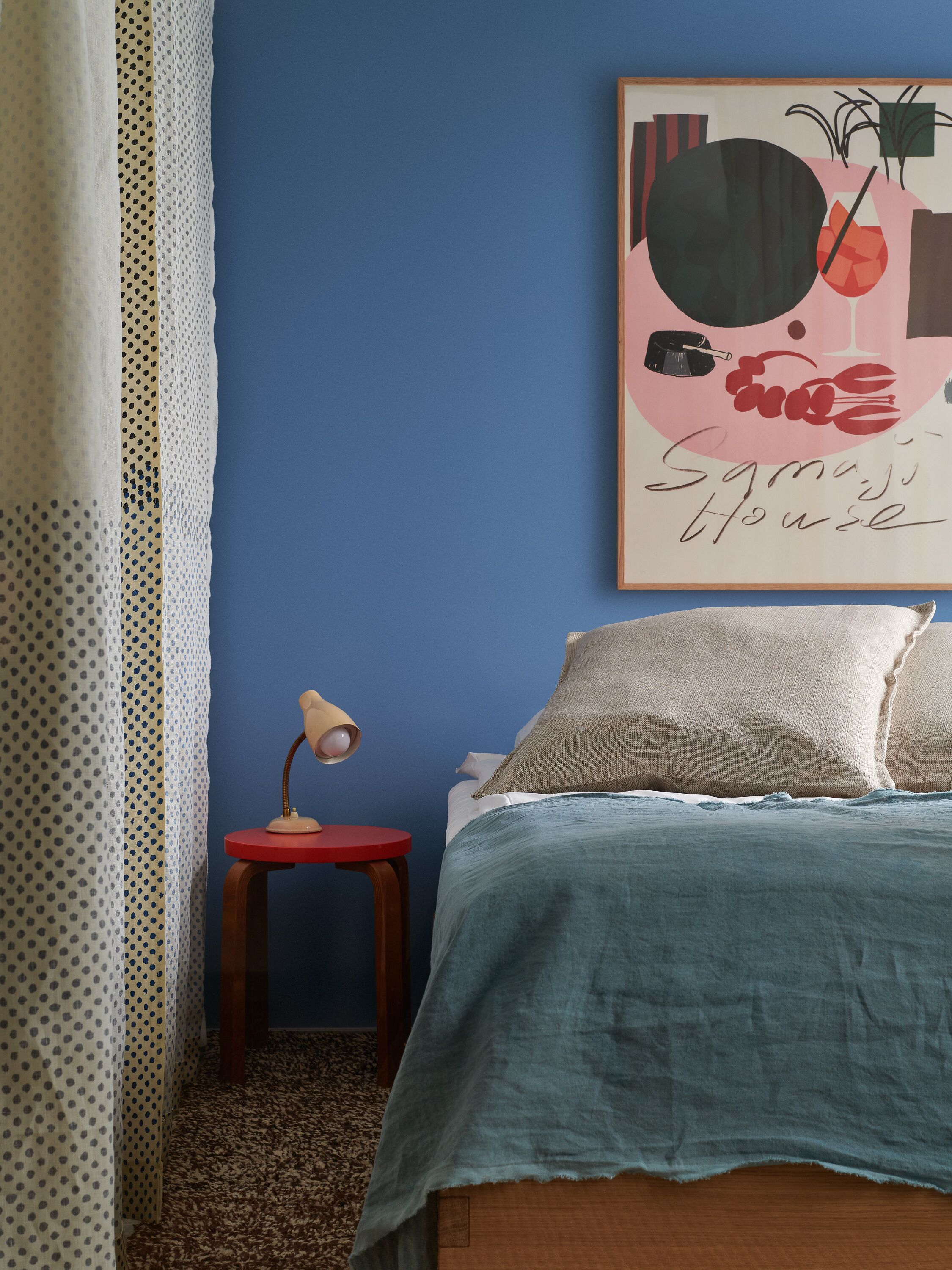 A bedroom with a blue wall, a bed with a blue bedspread, a red side table with a lamp, and a framed artwork on the wall.