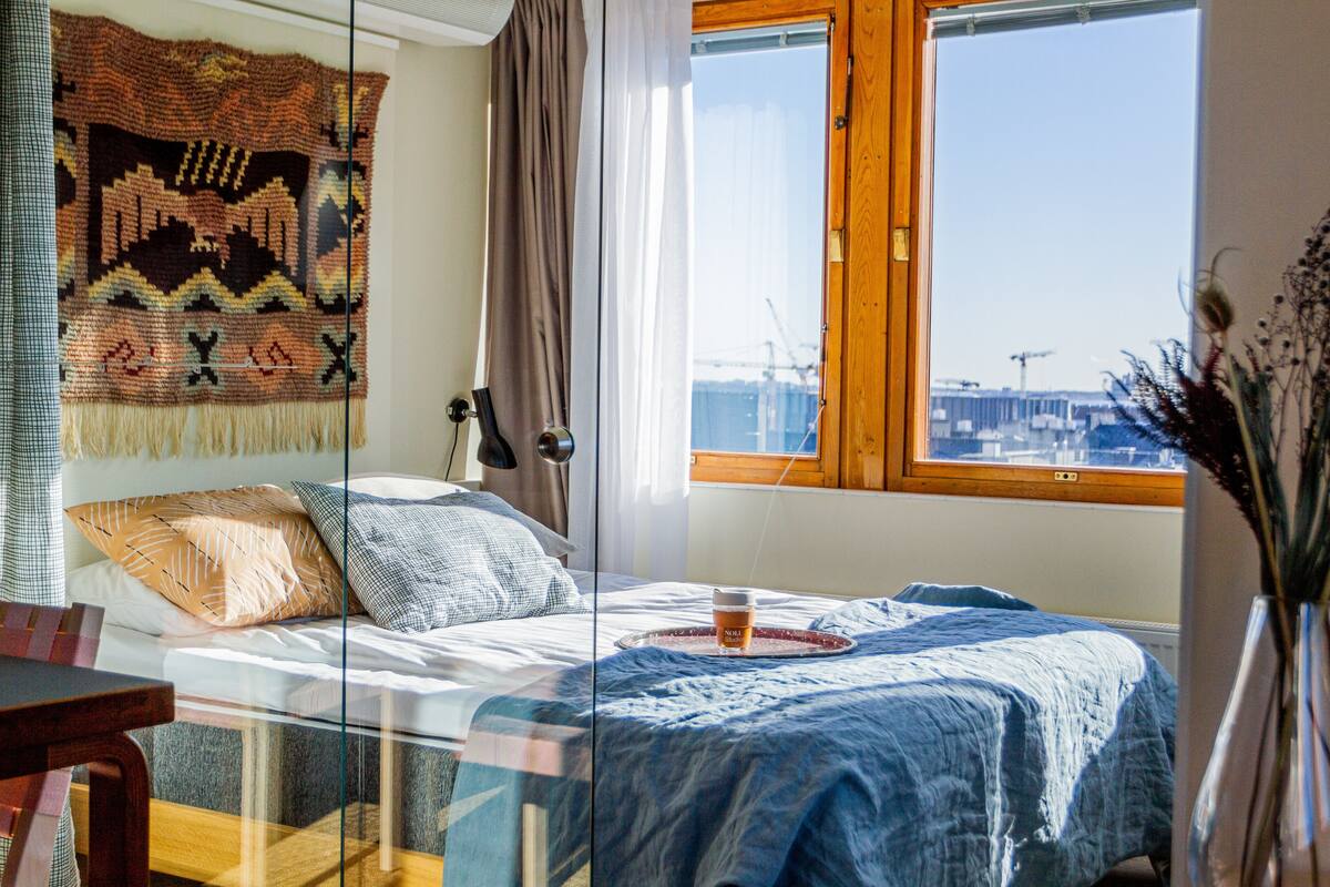 Une chambre à coucher avec un lit, une fenêtre donnant sur des immeubles, une couverture et une tasse posée sur le lit.