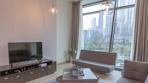 A modern living room with a large flat-screen TV, a grey sofa, a coffee table, and a city view through large windows. - Bravo Way Burj Vista 1 (Dubai)