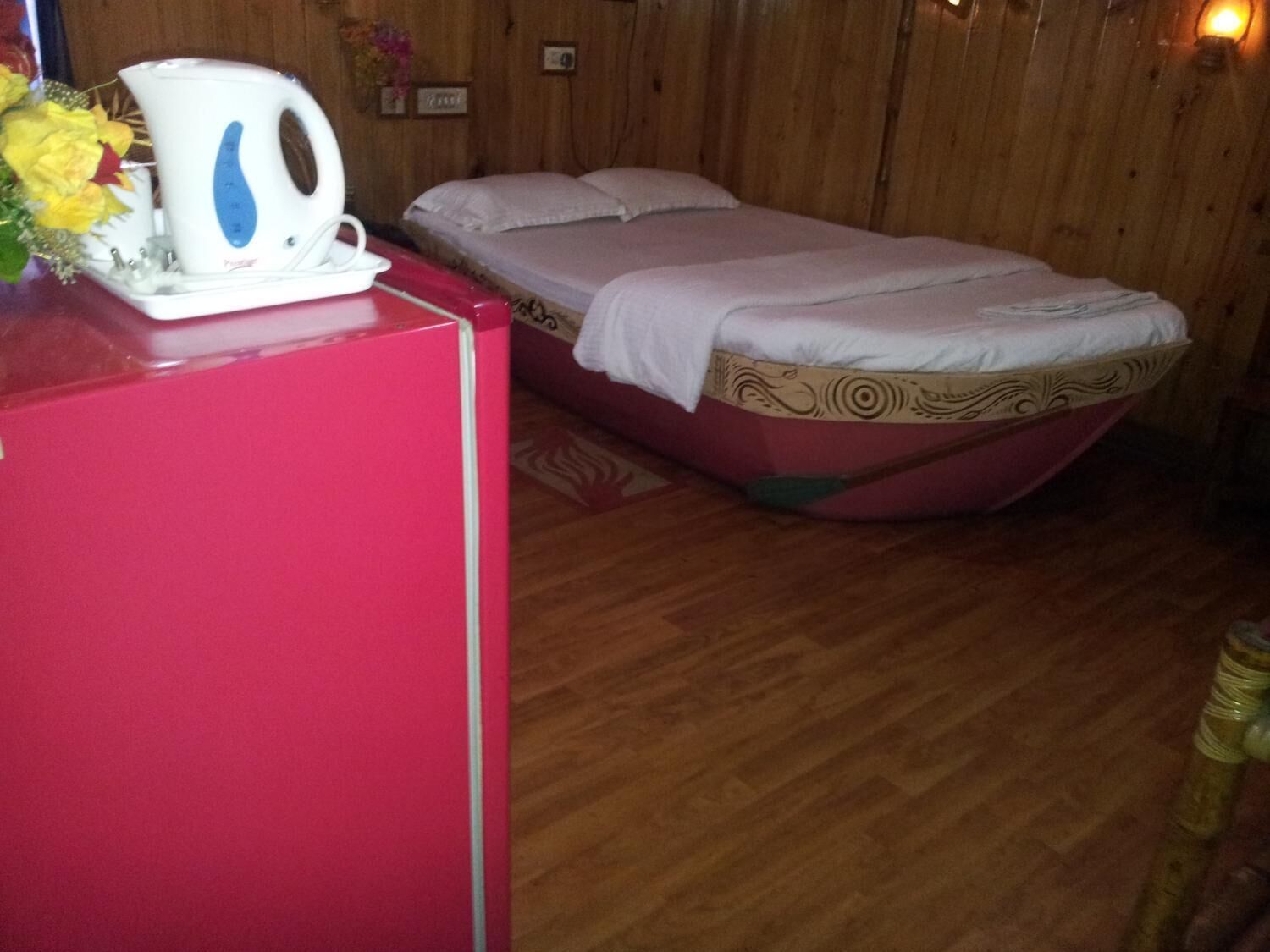 A room with a bed, a red refrigerator, and a kettle on a table.