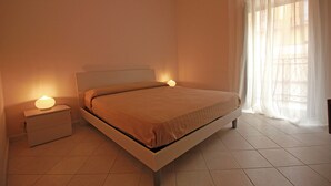 A bedroom with a bed, bedside lamps, a nightstand, and a window with sheer curtains.