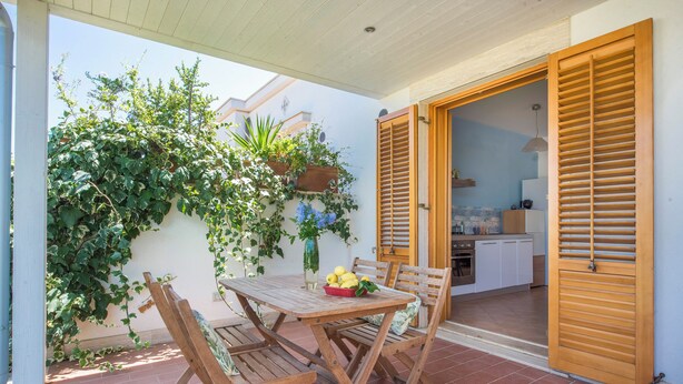 Un patio con una mesa y sillas de madera, una cesta de limones y una cocina visible a través de una puerta abierta.