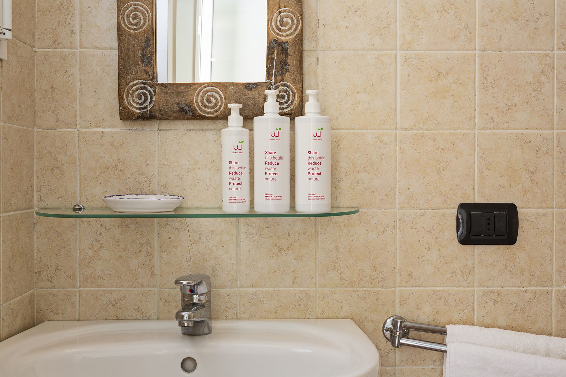 Um banheiro com pia, espelho e três frascos de shampoo orgânico em uma prateleira de vidro.