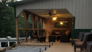 Eine überdachte Terrasse im Freien mit Holzboden und Sitzgelegenheiten, einem Deckenventilator und einem großen Fenster mit Blick auf einen bewaldeten Bereich.