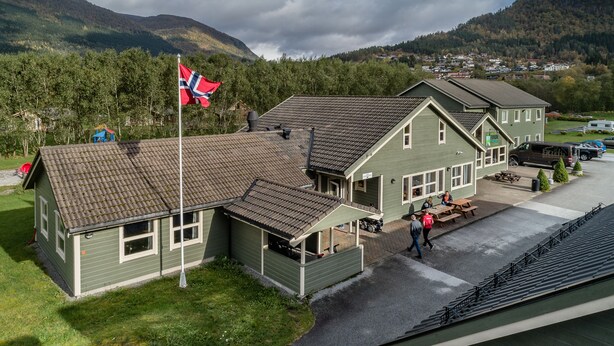 Un edificio de dos pisos con fachada verde y techo de tejas, que cuenta con un asta con bandera, una mesa de picnic y un pequeño aparcamiento.
