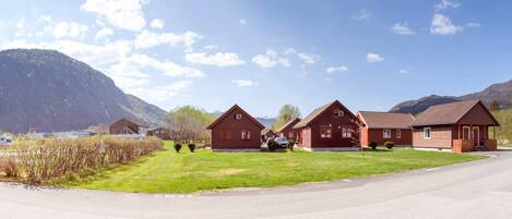 A row of red wooden cabins with a mountain backdrop, a clear sky, and a well-maintained lawn.