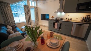 Una cocina moderna con microondas, estufa y lavavajillas. Hay una mesa de comedor con platos, vasos y un florero. Se ve un sofá y un televisor al fondo.