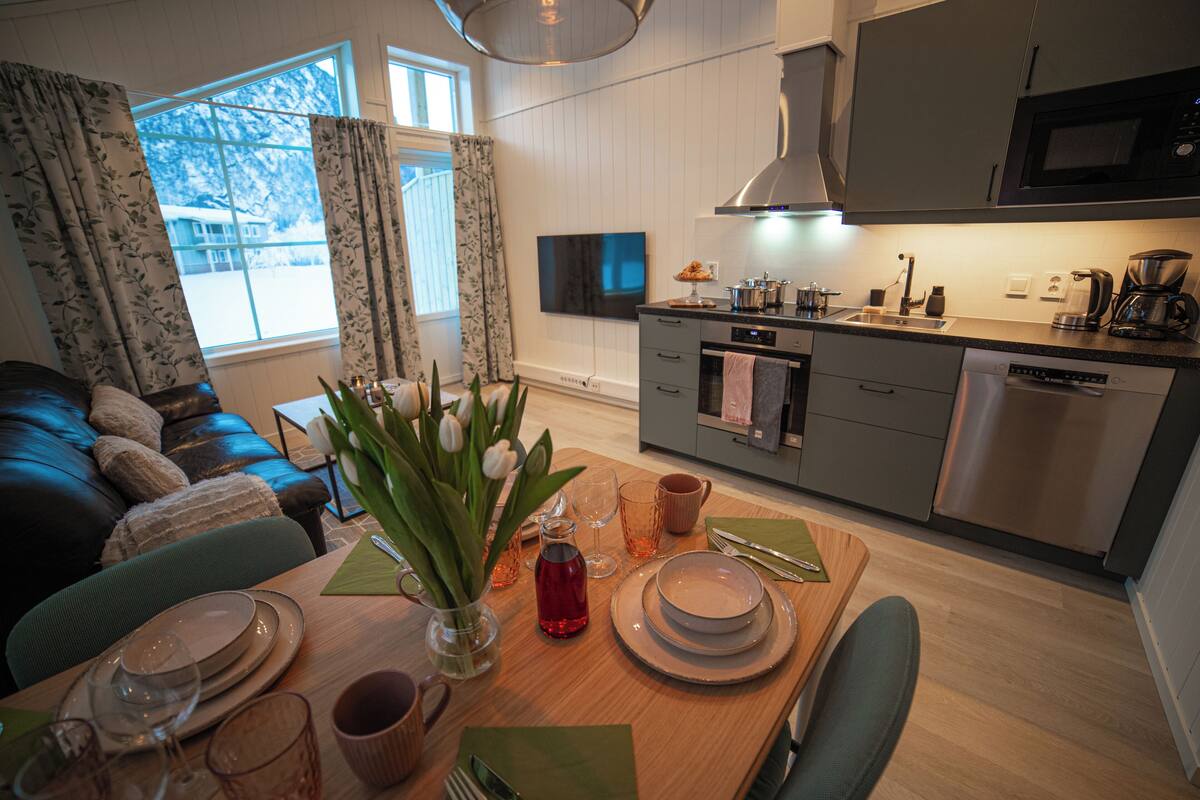 Eine moderne Küche mit Mikrowelle, Herd und Geschirrspüler. Ein Esstisch ist gedeckt mit Tellern, Gläsern und einer Blumenvase. Im Hintergrund sind eine Couch und ein Fernseher zu sehen.