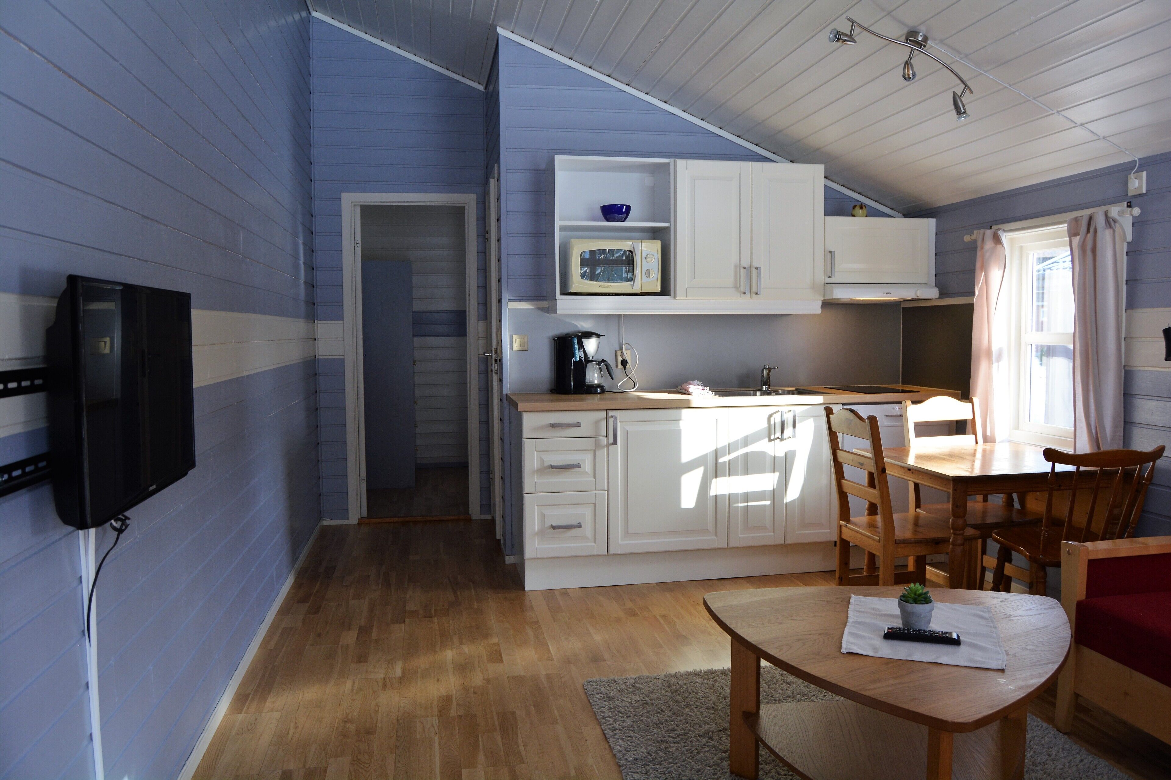 A compact kitchen with white cabinets, a microwave, and a coffee maker. There is a dining area with a table and chairs, and a television mounted on the wall.