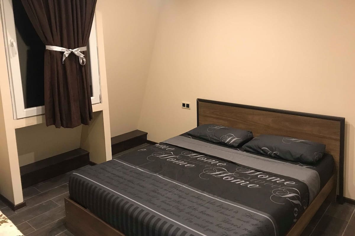 A bedroom with a bed, dark curtains, a window, and a bedside table.