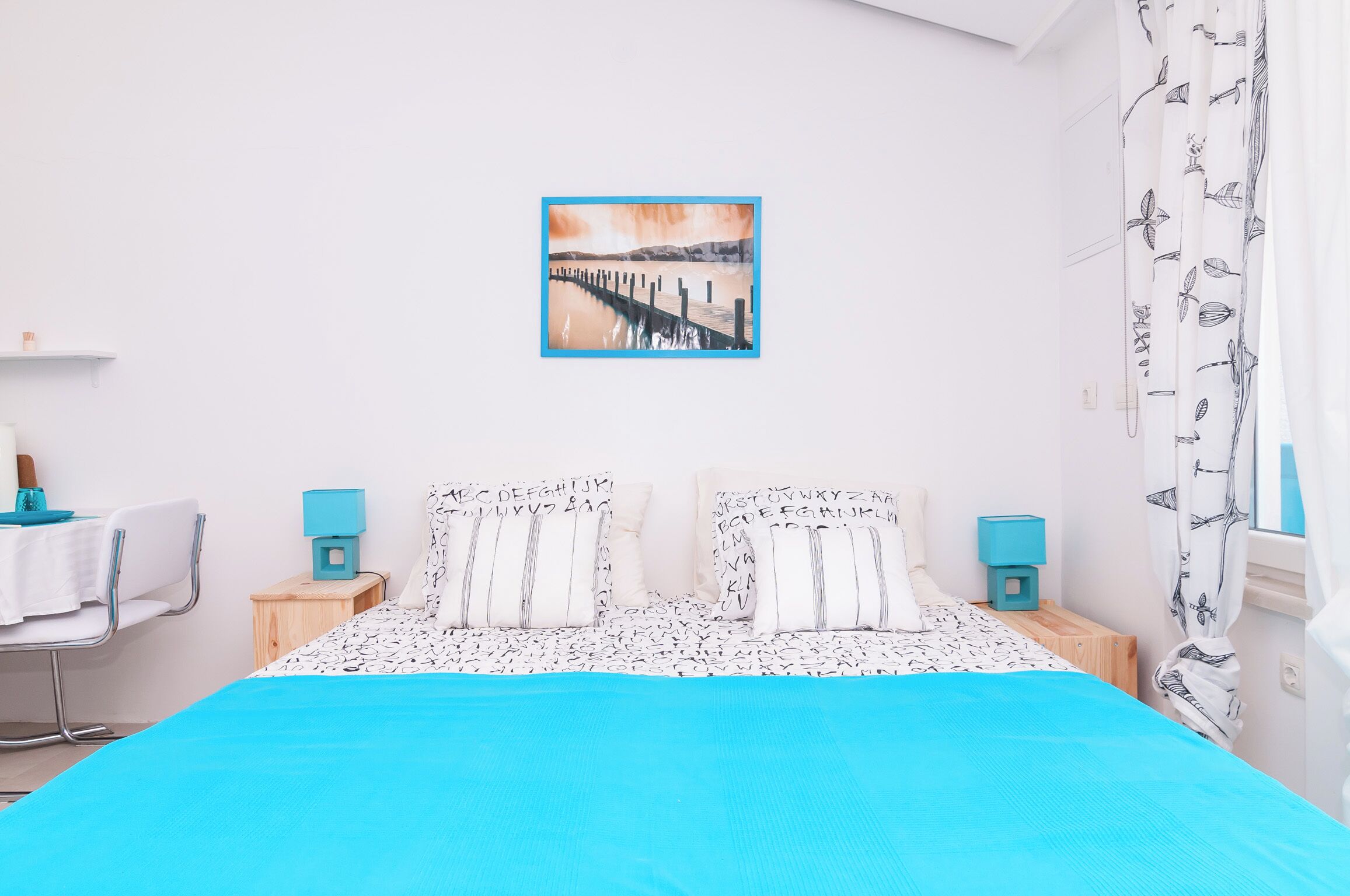 Quarto com cama, duas mesas de cabeceira, uma cadeira, um quadro na parede e janela com cortinas.