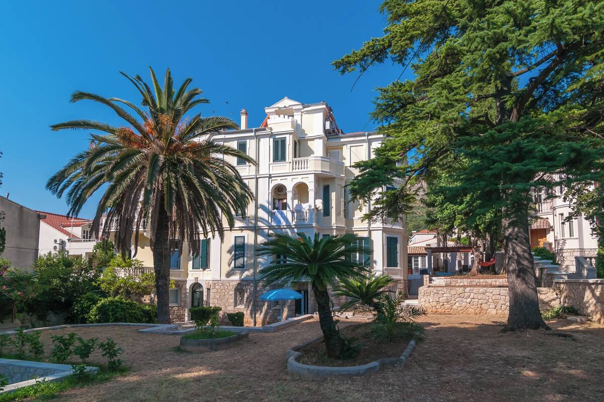 Un grand bâtiment historique doté de nombreux balcons et de fenêtres en arc de cercle, entouré d’un jardin aménagé avec des palmiers et un mur en pierre.