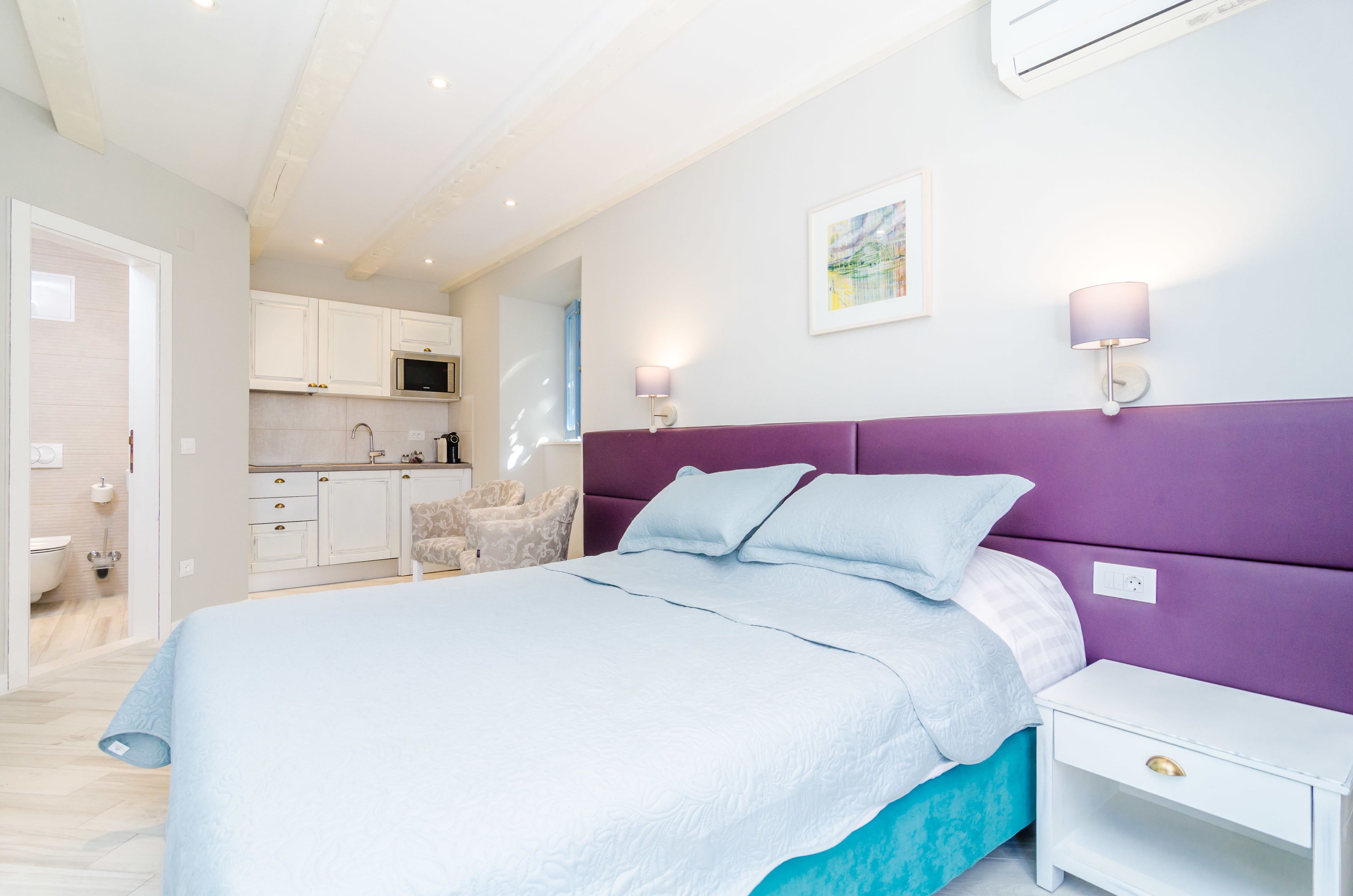 A bedroom with a bed, a nightstand, a chair, and a kitchenette in the background.