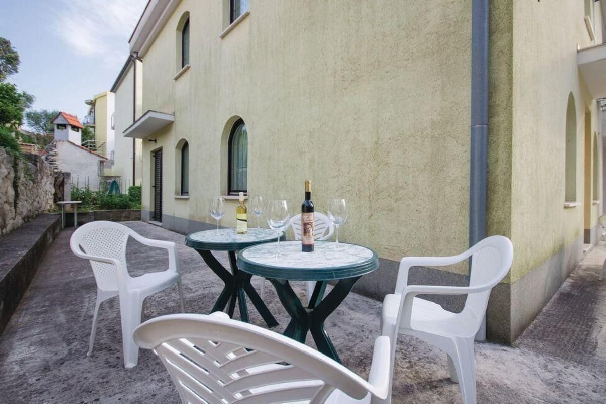 Um pátio externo com duas mesas redondas, cada uma com uma garrafa de vinho e copos, e cadeiras de plástico branco.
