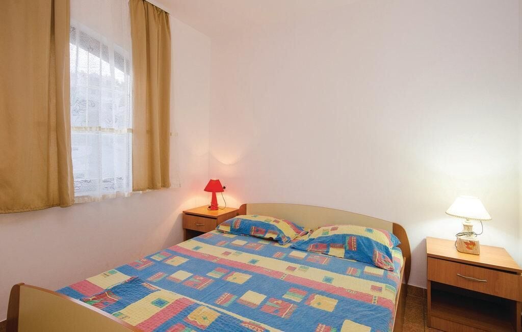 A single bed with a colorful quilt, two bedside tables with lamps, a window with curtains, and a plain wall.