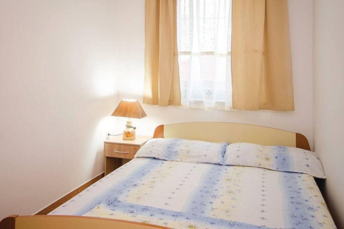 Uma cama de solteiro com colcha florida, uma mesa de cabeceira com abajur e uma janela com cortinas.