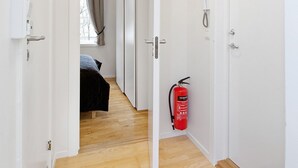 Un couloir doté d’un plancher en bois, de murs blancs et d’une porte donnant sur une chambre à coucher avec un lit et des rideaux. Un extincteur est fixé au mur.