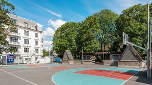 Un terrain de basket en plein air, doté d’une surface bleue et rouge, entouré d’arbres et bordé d’un bâtiment à la façade blanche.