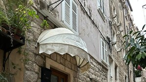 Un edificio de piedra con puerta de madera y un paraguas blanco. Hay escalones que conducen a la entrada, y se ve una planta en maceta en el suelo.