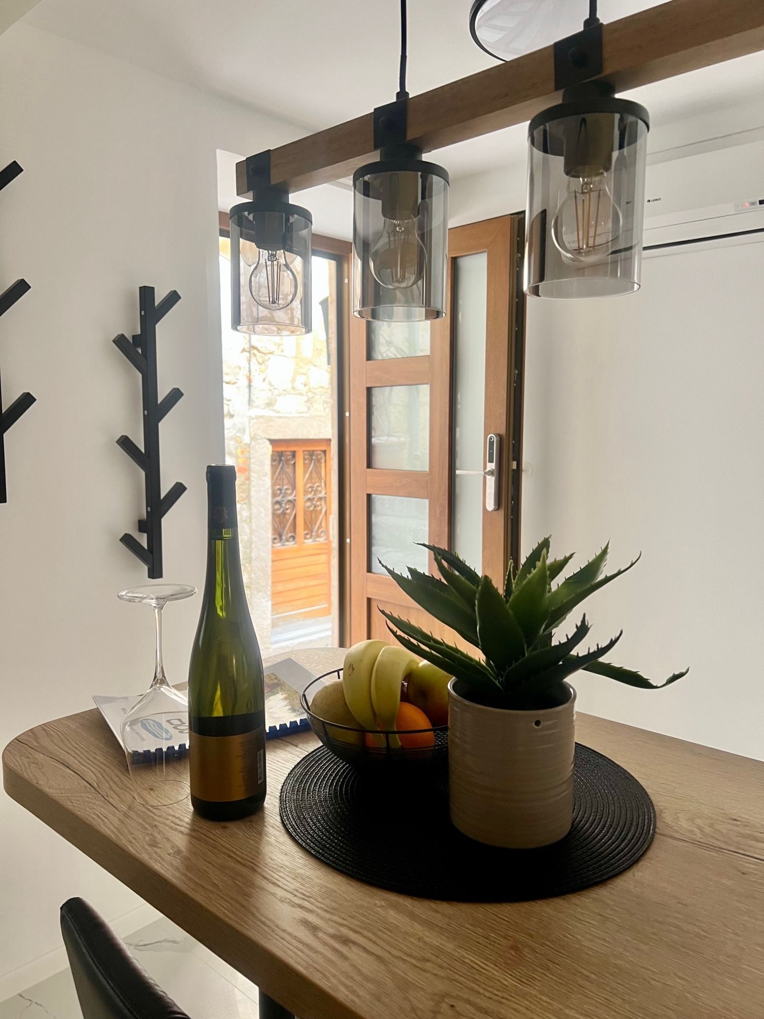 Una mesa de comedor con una botella de vino, una copa y un frutero. Hay una planta en maceta sobre la mesa.
