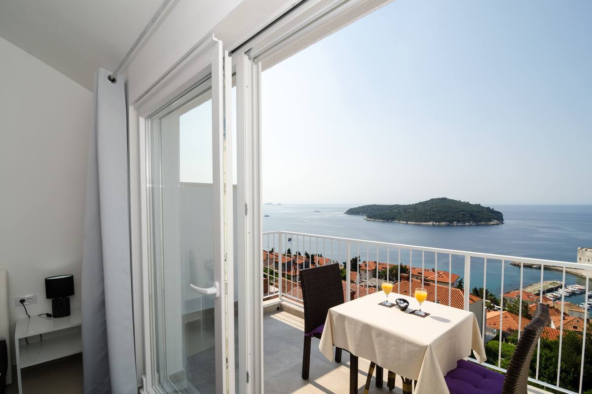 A balcony with a table set for two, overlooking a coastal view with a small island and a marina.
