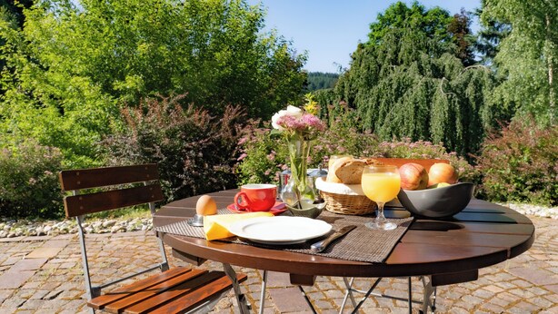 Ein runder Holztisch draußen mit einem Frühstücksbuffet, darunter Brot, Obst und ein Glas Orangensaft.