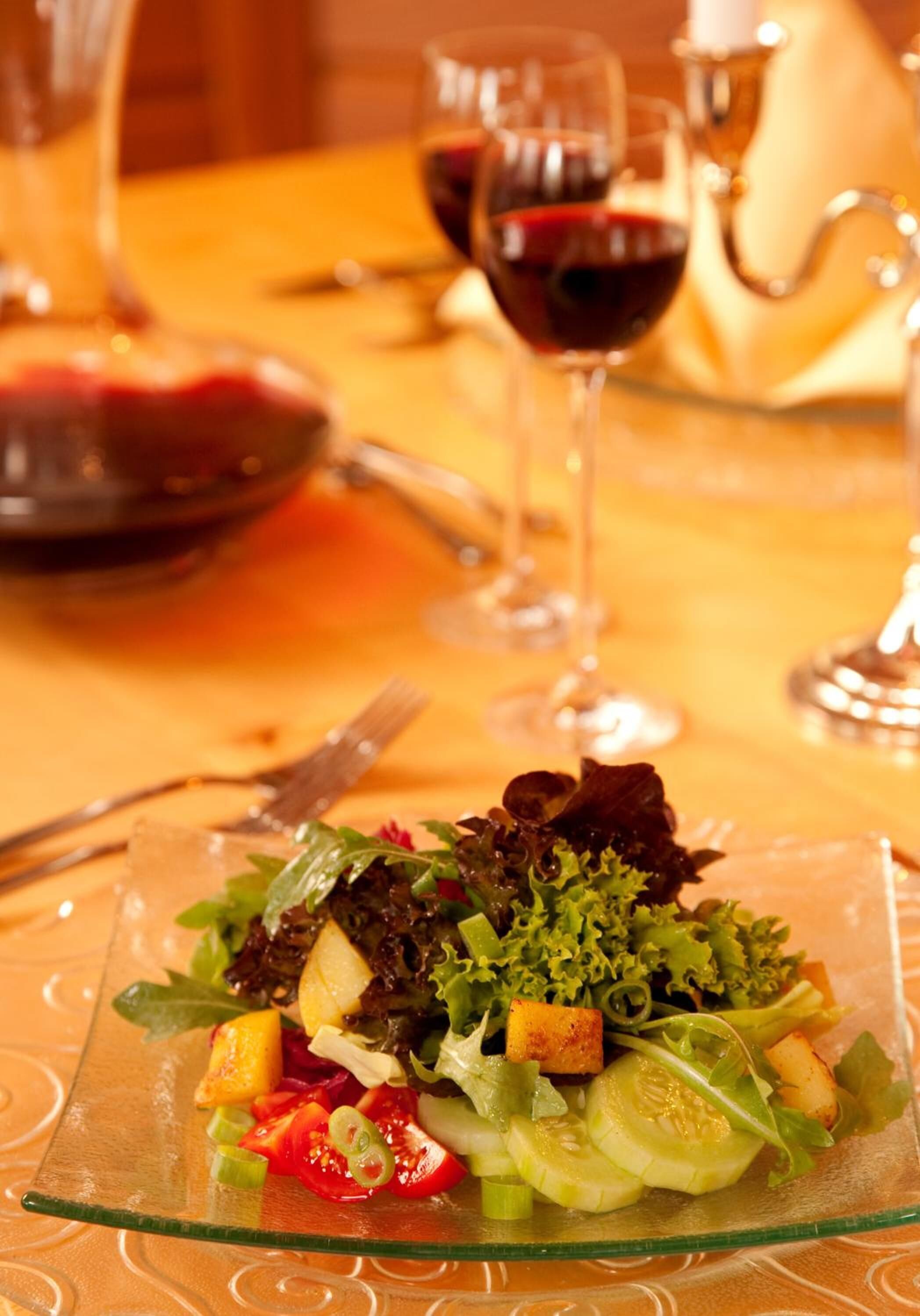 A glass plate with a salad on a table, accompanied by wine glasses and a teapot.