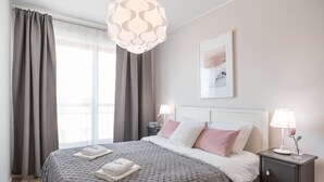 Ein ordentlich bezogenes Bett mit einer gemusterten Tagesdecke, ein Nachttisch mit Lampe und einer Vase sowie ein gerahmtes Bild an der Wand.
