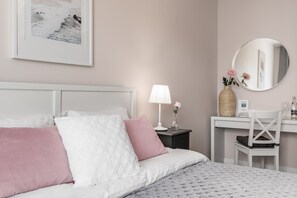 A neatly arranged bedroom with a bed, a bedside table with a lamp, a mirror, and a desk with a chair.