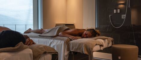 Une salle de spa moderne équipée d’une table de massage, d’un fauteuil de massage séparé et d’une grande fenêtre laissant entrer la lumière naturelle.