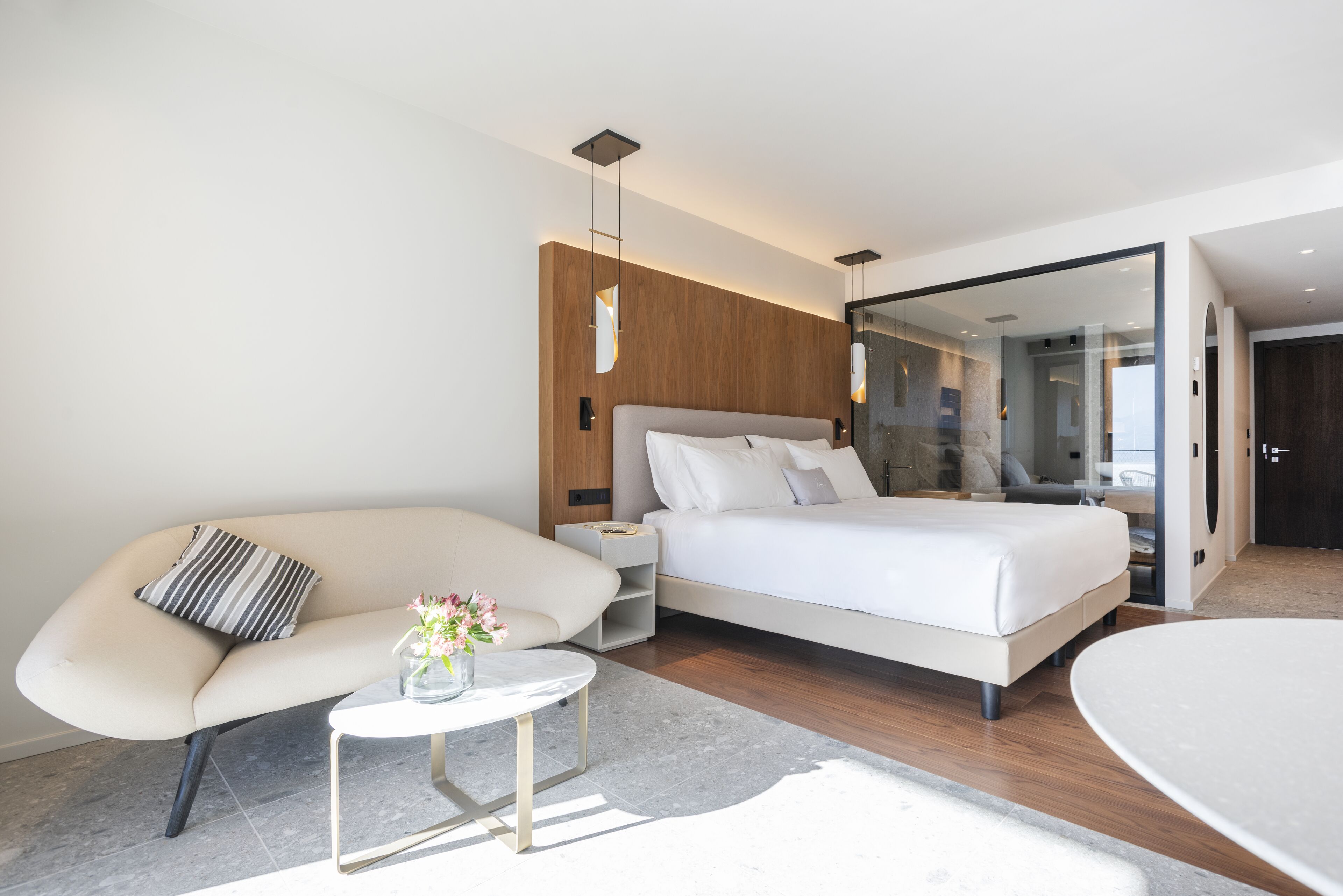 Un dormitorio moderno con una cama, un sofá, una mesita con un florero y un tabique de vidrio.