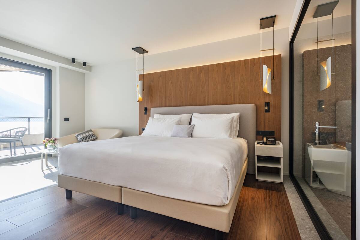 Un dormitorio moderno con una cama grande, mesitas de noche, cabina de ducha de vidrio y un balcón con mobiliario exterior.