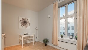 Ein modernes, minimalistisches Zimmer mit Holzboden, einem weißen Schreibtisch mit zwei Stühlen, einer Topfpflanze und einem Fenster mit beige farbenen Vorhängen.