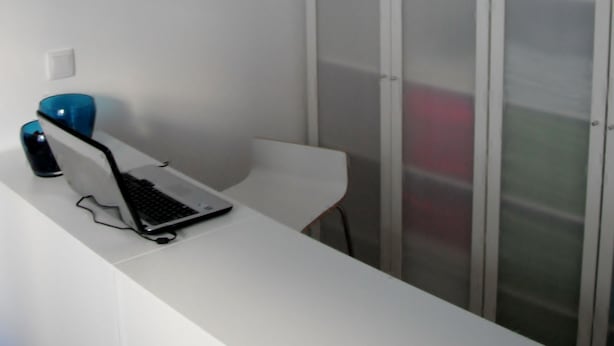 A modern room with a white desk, a laptop, and a blue vase. There is a white chair and a white wardrobe with frosted glass doors. - 70's Hostel (Mafra)