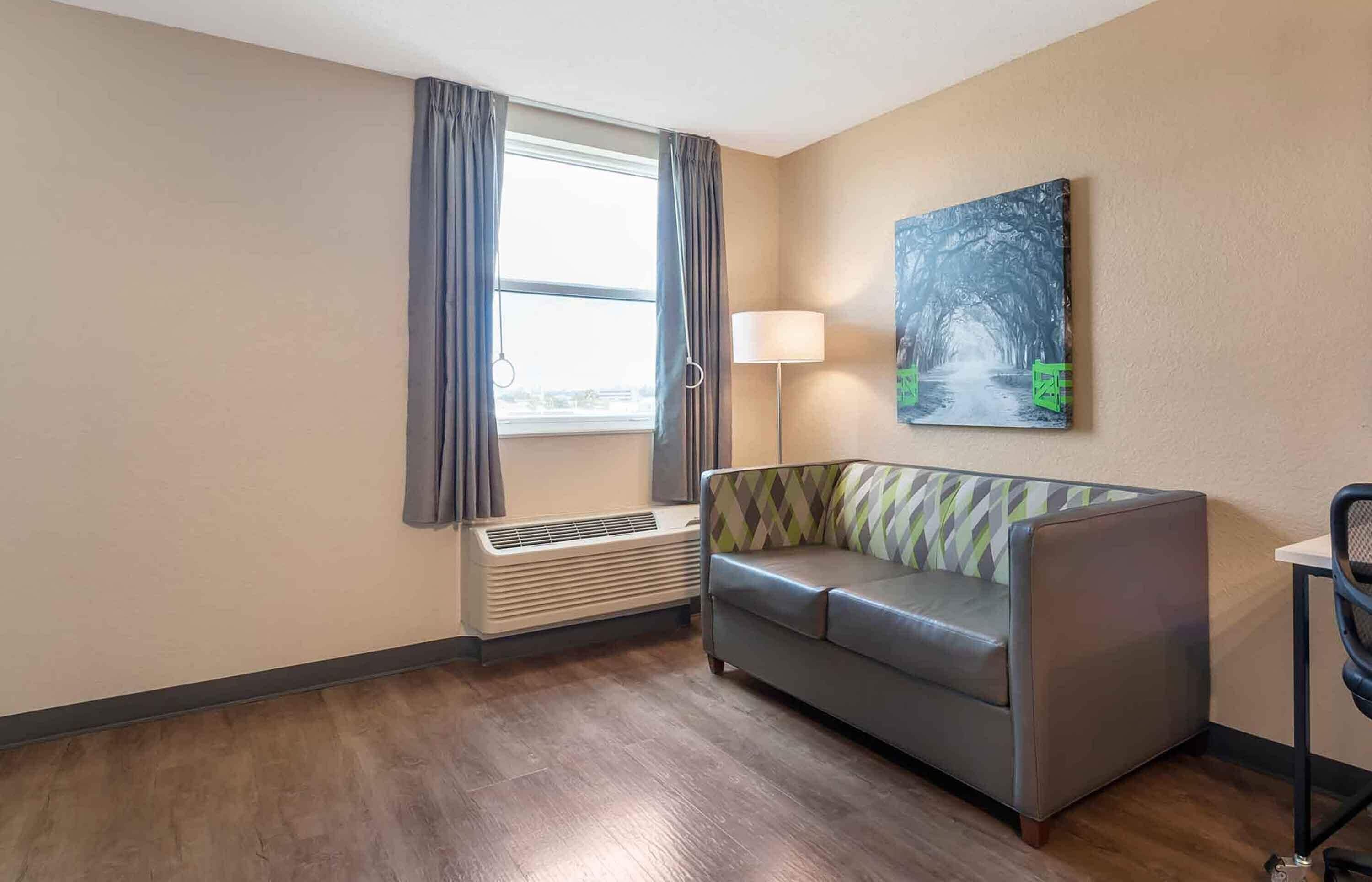 A hotel room with a sofa, a standing lamp, a painting, and a window with curtains.