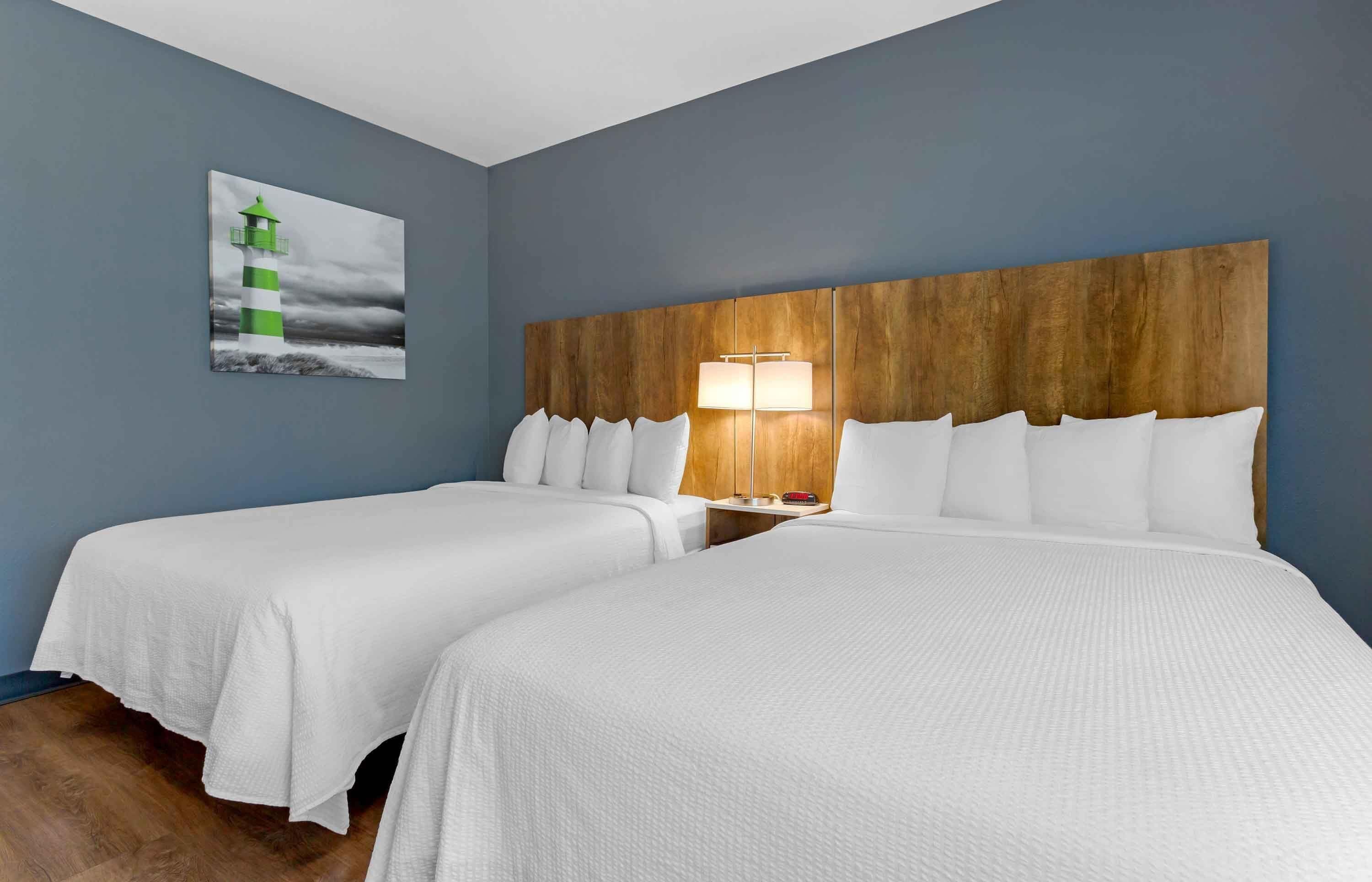 Habitación de hotel con dos camas, cabecera de madera, mesita de noche con lámpara y un cuadro de un faro.