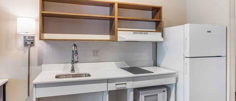 Una cocina compacta con encimera blanca, microondas integrado, refrigerador blanco y estantes de madera sobre la encimera.