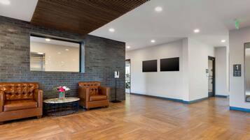 A spacious lobby with a wooden floor, two leather armchairs, a round table with a vase, a large mirror, and a modern ceiling design.