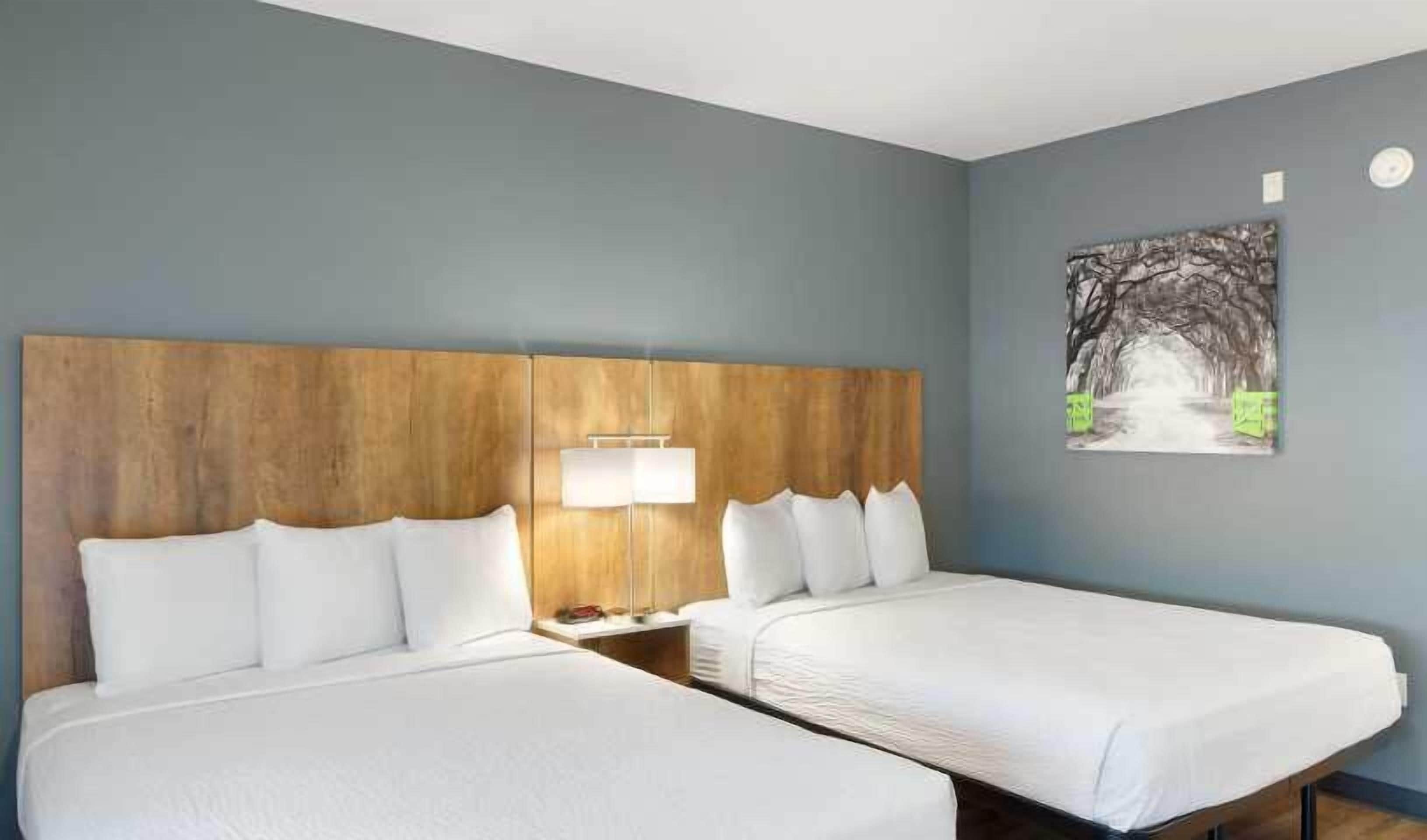 Ein Hotelzimmer mit zwei Betten, einem Holz-Kopfteil, einem Nachttisch mit Lampe und einem Bild an der Wand.