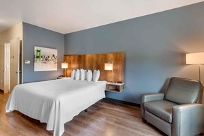 A hotel room with a bed, a headboard, a chair, a nightstand, and a wall-mounted lamp.