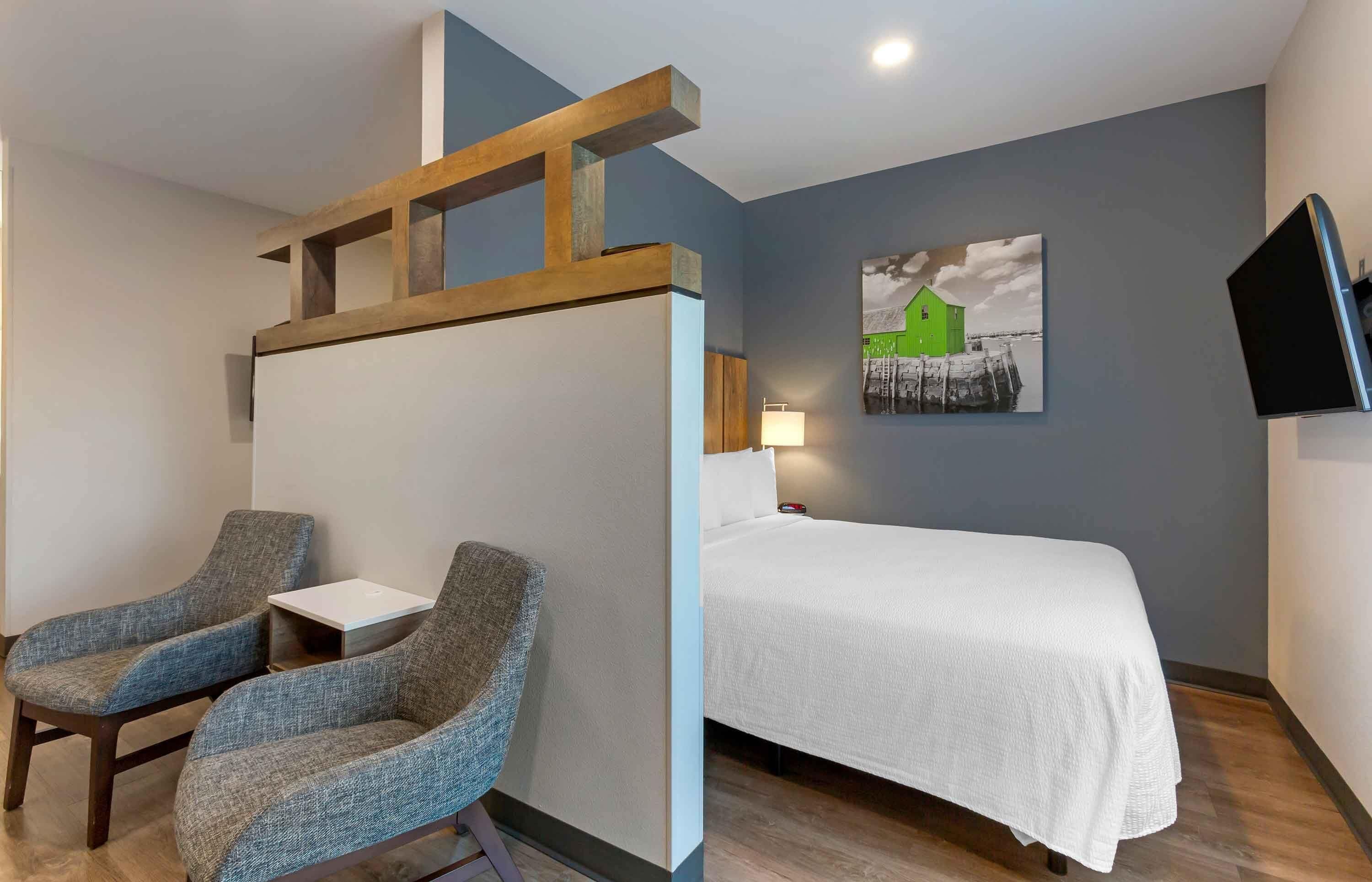 A hotel room with a bed, two armchairs, a small table, a painting, and a TV.