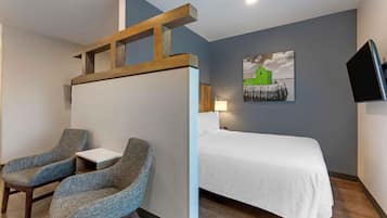 A hotel room with a bed, two armchairs, a small table, a painting, and a TV.