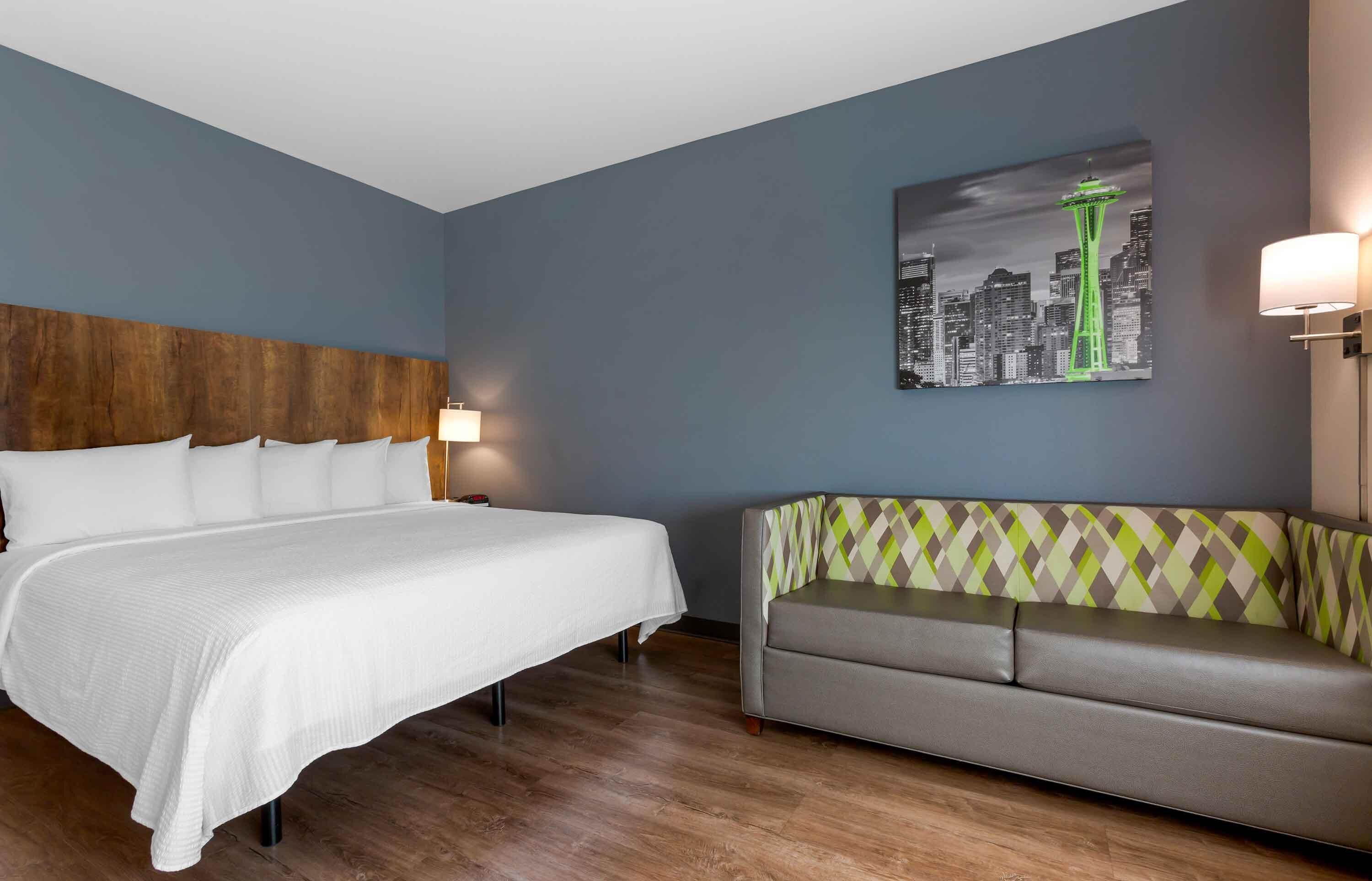 A hotel room with a bed, a sofa, a cityscape painting, and a lamp.