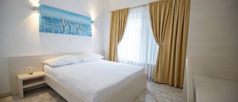 Habitación de hotel con cama, mesita de noche, lámpara de pared, un cuadro y ventana con cortinas.
