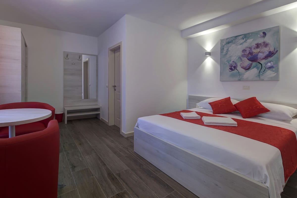 A hotel room with a bed, a red chair, a small table, and a painting on the wall.