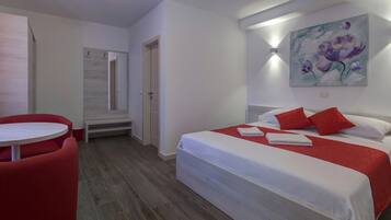 A hotel room with a bed, a red chair, a small table, and a painting on the wall.