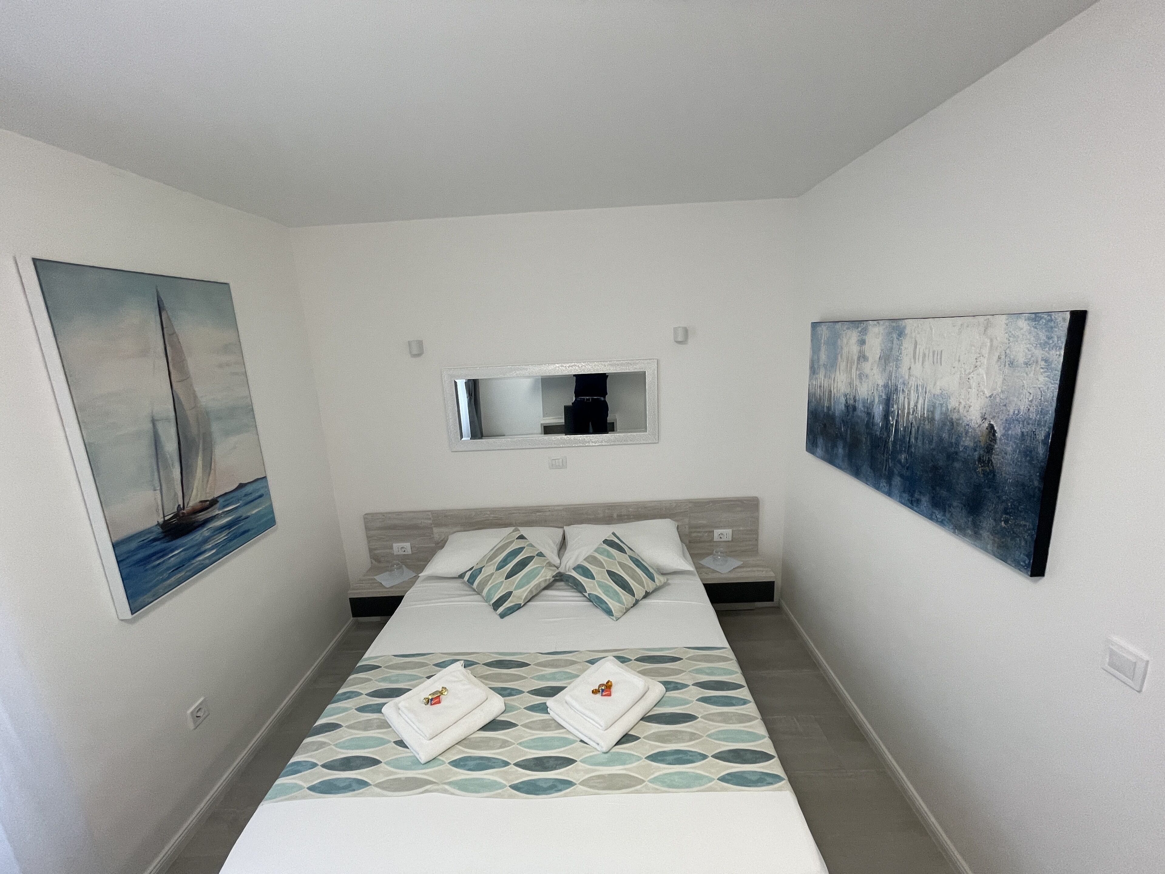 Une chambre d’hôtel avec un lit, un tableau représentant un voilier, un miroir et un tableau représentant un paysage urbain.