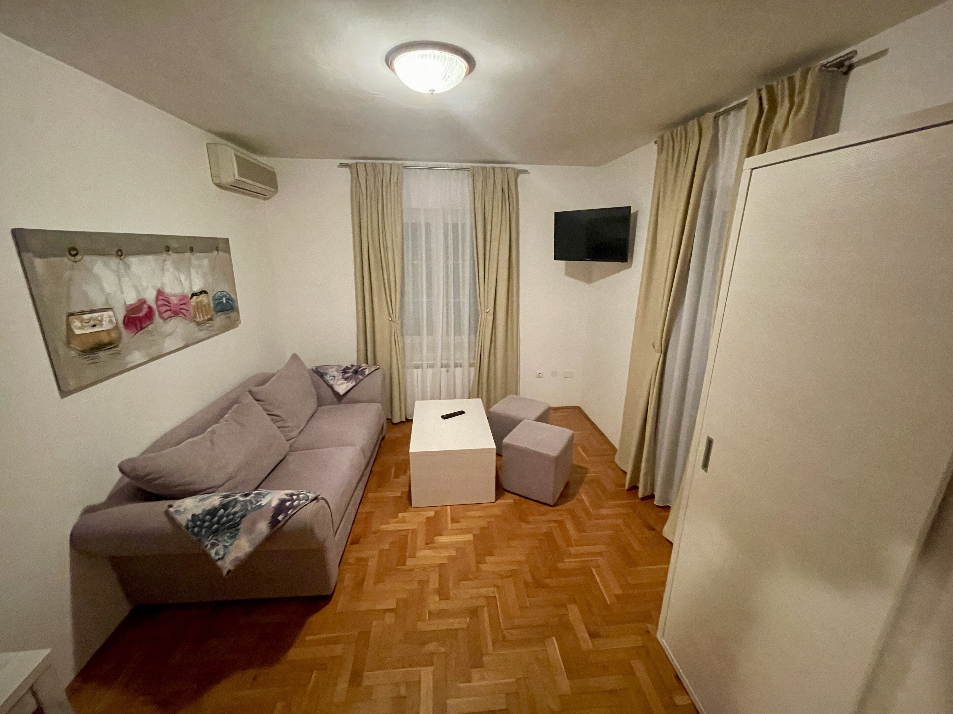 Una habitación pequeña y bien iluminada con piso de madera en espiga, un sofá gris, una mesita de centro pequeña y un televisor montado en la pared.
