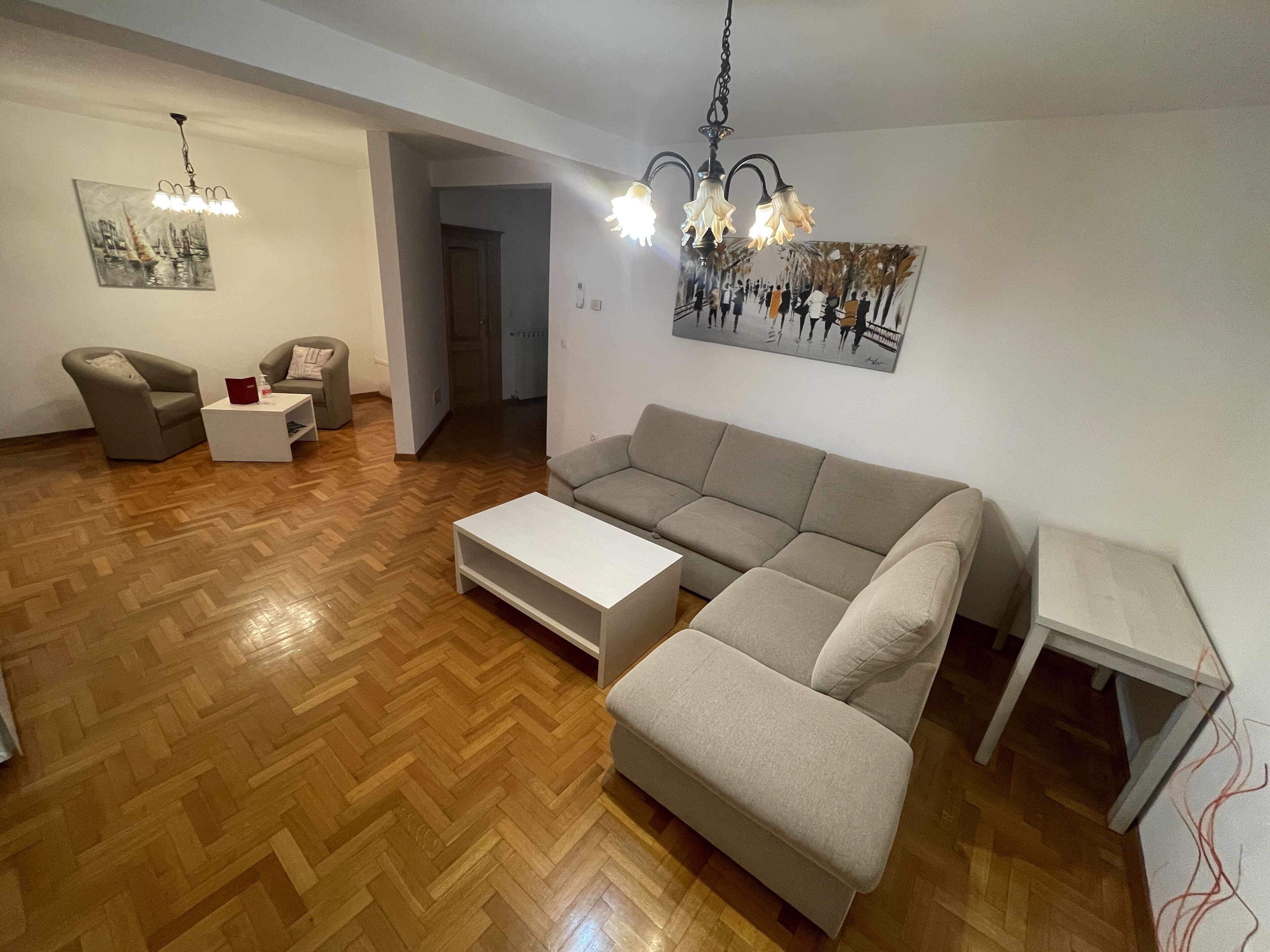 Un salón con suelo de madera en patrón de espiga, un sofá gris, una mesa de centro blanca, un sillón gris y una mesita auxiliar blanca. Hay un cuadro en la pared y una araña de techo sobre el sofá.