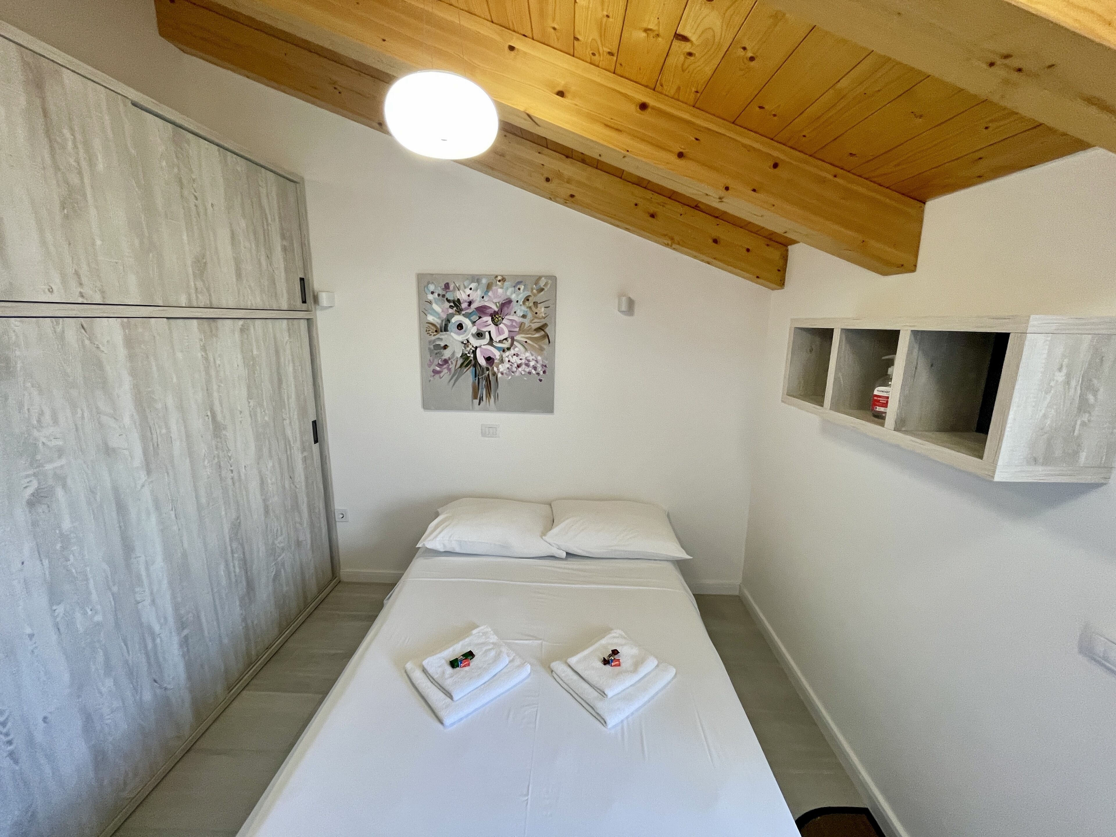 A bedroom with a wooden ceiling, a bed with white linens, a floral artwork on the wall, and a small shelf with a bottle.